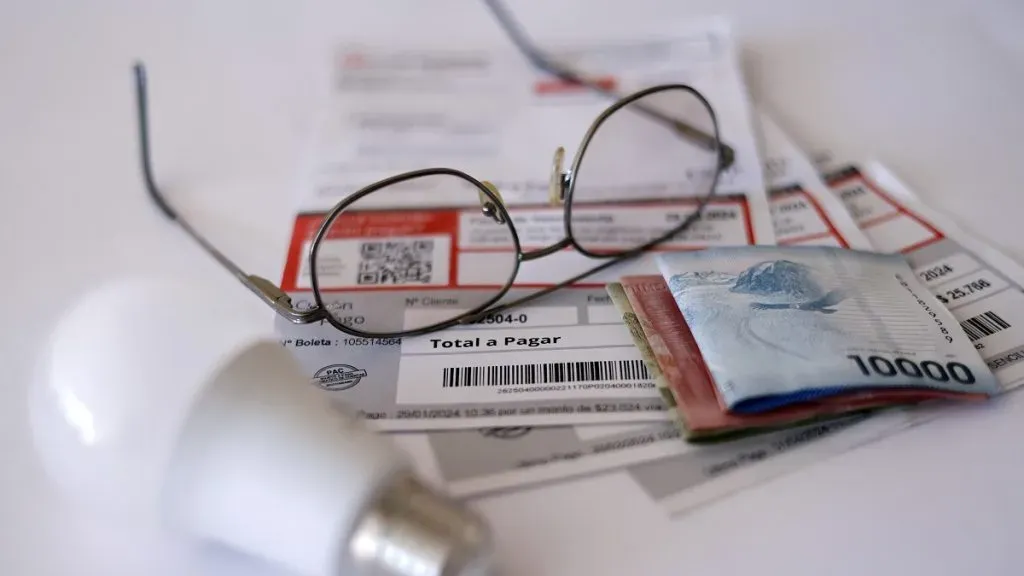 Sobre una mesa hay dos cuentas de luz; billetes de mil, cinco mil y 10 mil; unos lentas; y una ampolleta blanca.