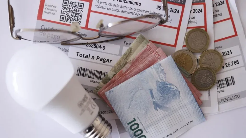 En una mesa blanca hay cuentas de la luz, billetes, unos lentes y una ampolleta.