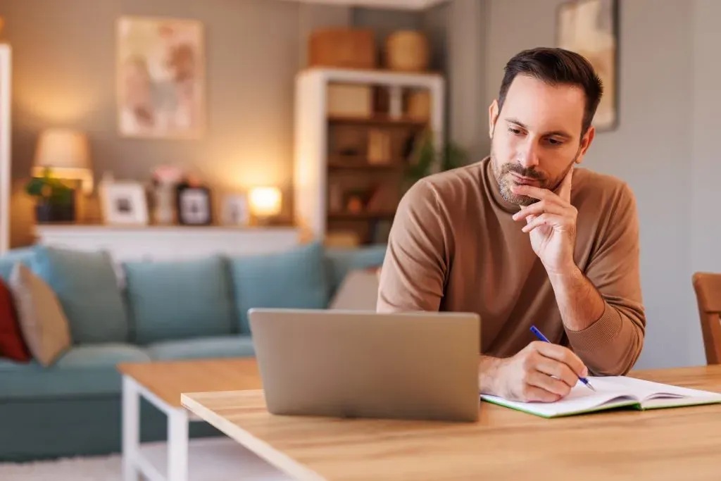 Portrait of focused businessman writing in diary while doing online research over computer in home office