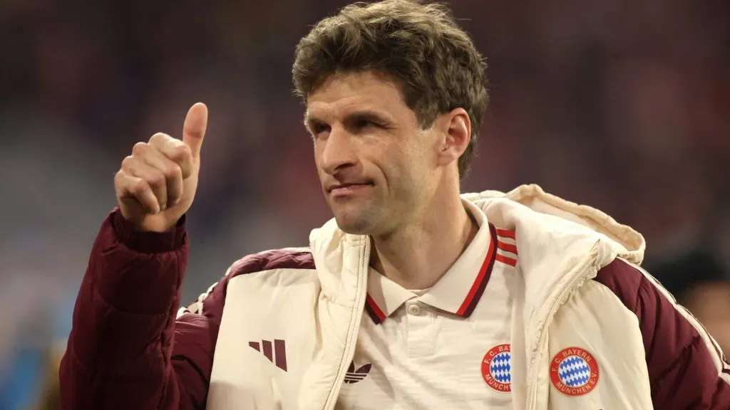 Thomas Müller, com a jaqueta bege e vinho do Bayern de Munique, em campo, acenando com a mão