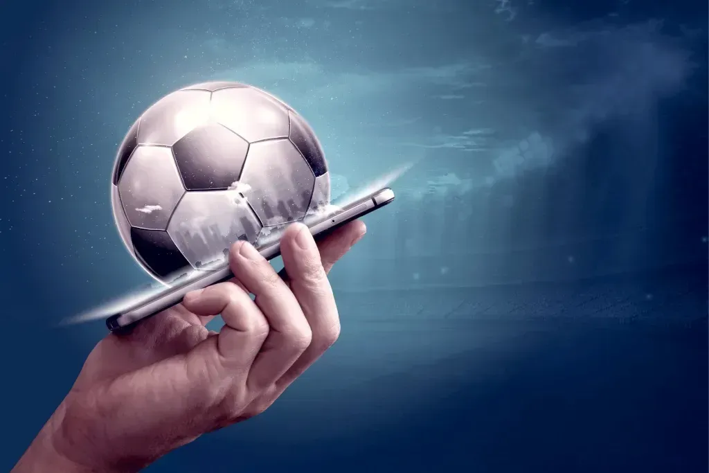 Com um fundo em tons de azul, uma mão segura um telemóvel, do qual emerge uma bola de futebol em 3D.