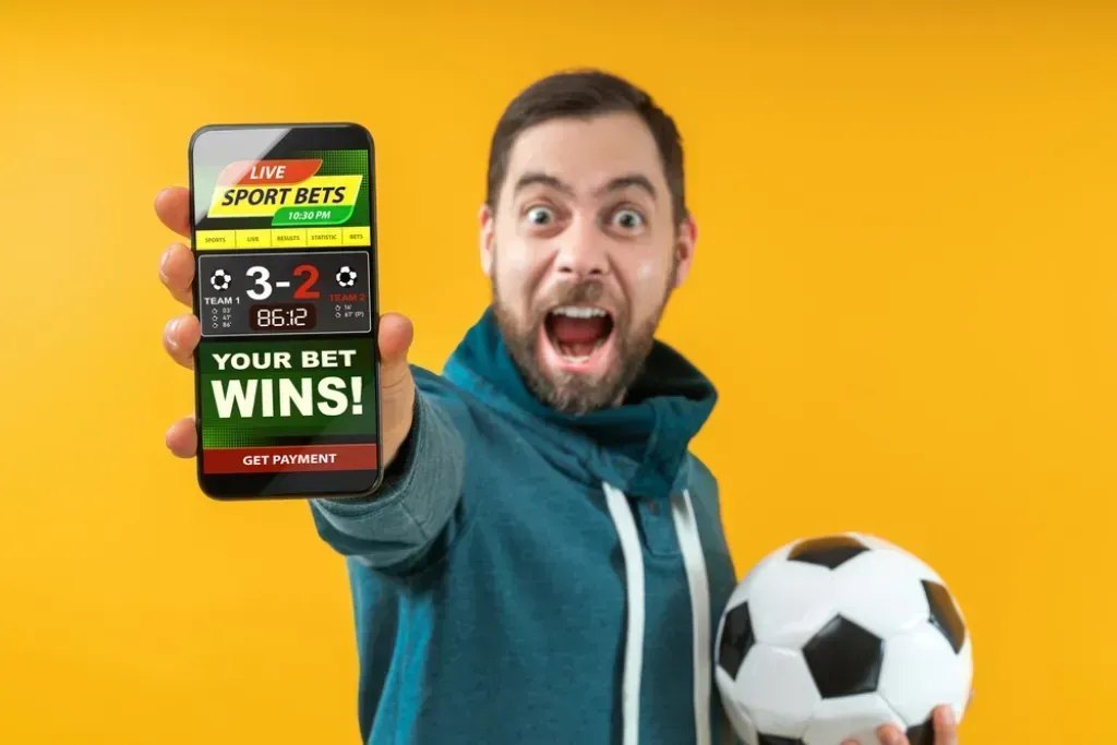 Sobre um fundo amarelo, um apostador segura uma bola de futebol na mão esquerda, enquanto estica o braço direito para mostrar o écran do telemóvel onde acompanha apostas ao vivo.